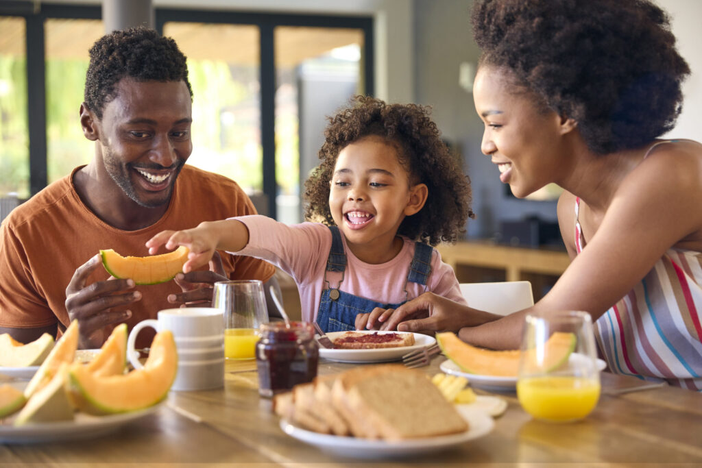 Family Shot With Parents And Daughter At Home Having Breakfast Spreading Jam On Bread At Table