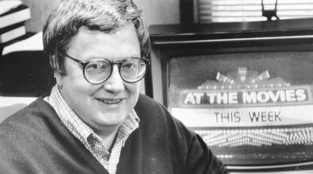 Black-and-white Roger Ebert in front of theater sign saying "At the Movies" in lights