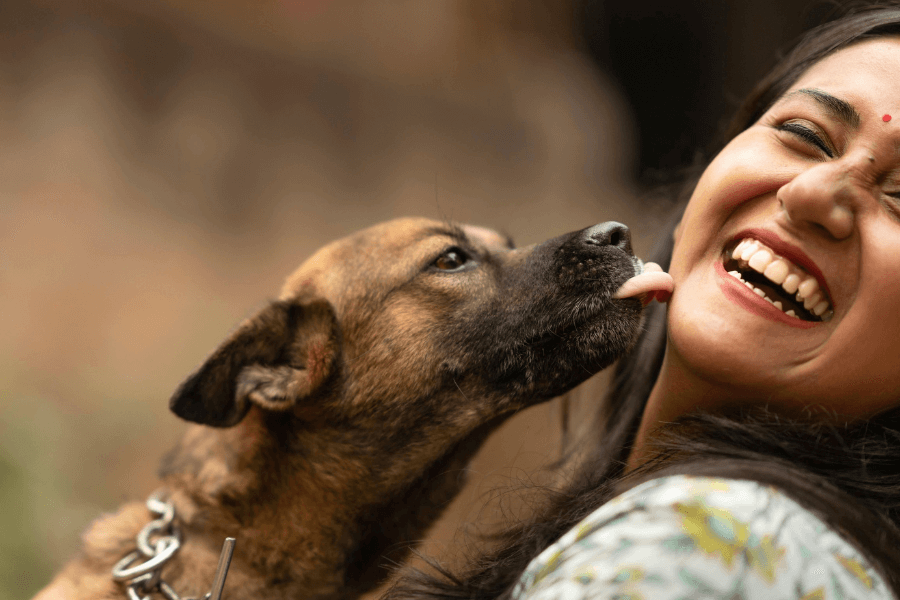 This heartwarming image captures a joyful moment between a woman and a dog. The dog, a light brown mixed breed, is affectionately licking the woman's face, while she laughs brightly with her eyes closed. The background is softly blurred, keeping the focus on the emotional connection between them.