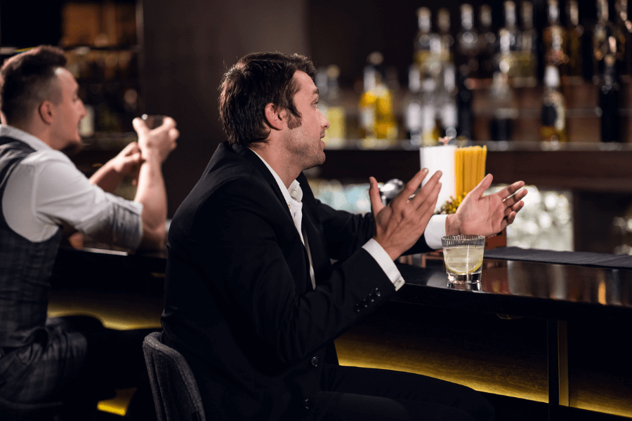 A man in a black suit gestures animatedly while sitting at a bar, holding a glass of liquor on the counter. Another man in a gray vest sits beside him, appearing to listen, with shelves of alcohol bottles blurred in the background.