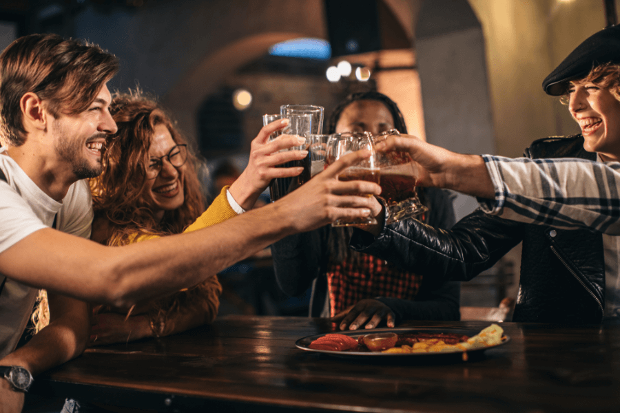 A group of friends cheers with glasses of beer at a wooden table in a lively bar or restaurant. They are smiling and laughing, with a plate of snacks, including fries and dipping sauces, visible on the table.