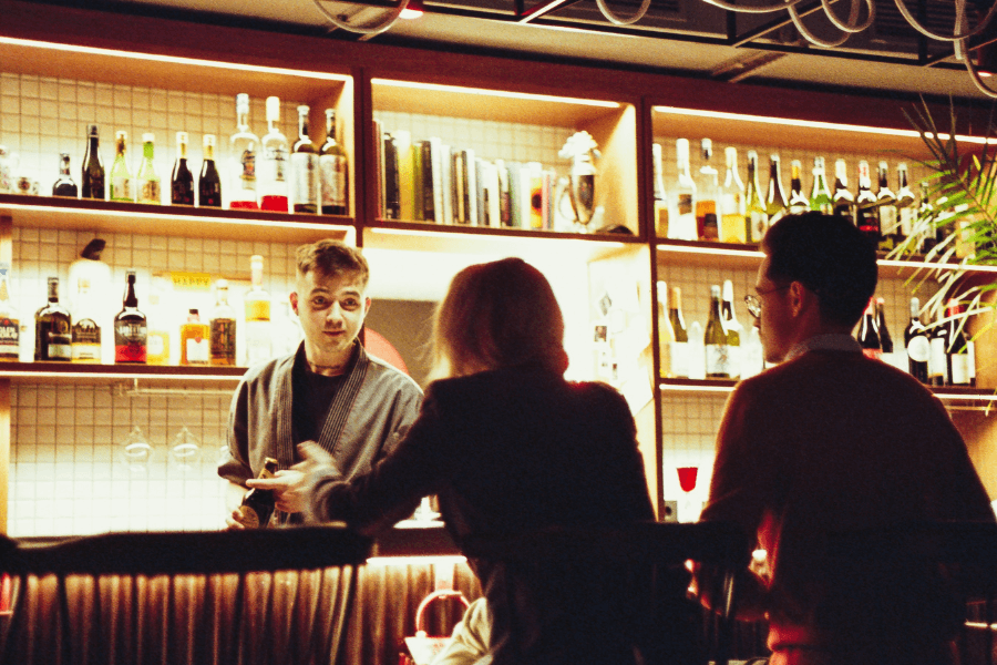 A bartender in a cozy, warmly lit bar interacts with two patrons sitting at the counter. Shelves in the background are stocked with bottles of liquor and wine, along with decorative items and plants.