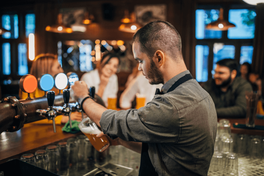 A bartender pours beer from a tap into a glass at a cozy bar, with customers smiling and chatting in the background. The warm lighting and wooden decor create an inviting atmosphere.