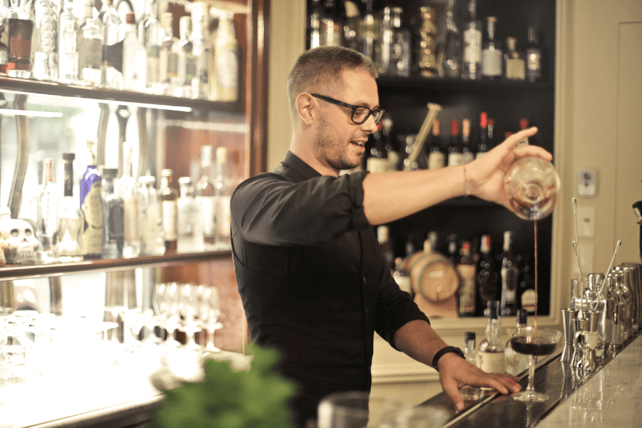 A bartender wearing glasses and a black shirt pours a cocktail from a mixing glass into a coupe glass at a well-stocked bar. Shelves behind him are lined with bottles and bar tools, adding to the elegant atmosphere.