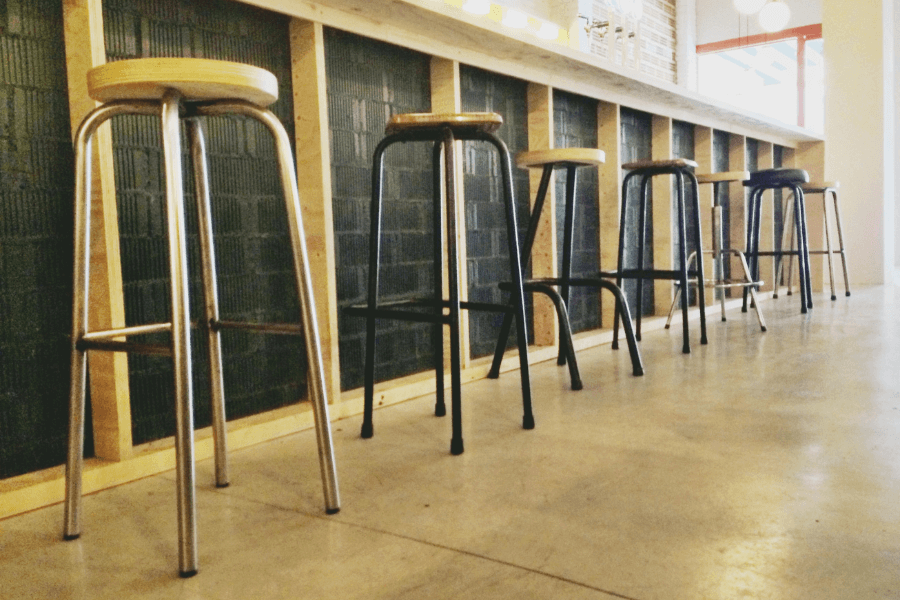 A row of metal and wood bar stools lined up against a wooden bar with a textured dark panel backdrop. The setting features a polished concrete floor and bright natural light from nearby windows.