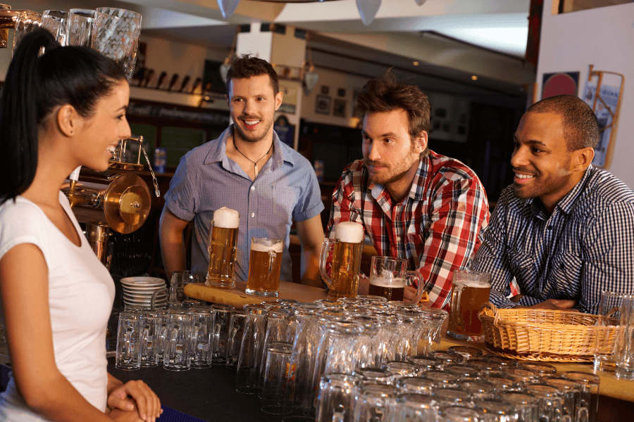 Three men sitting at a bar, each with a glass of beer in front of them, smile and chat with a bartender in a white shirt. The bar is brightly lit with rows of glassware and brewing equipment visible in the background.