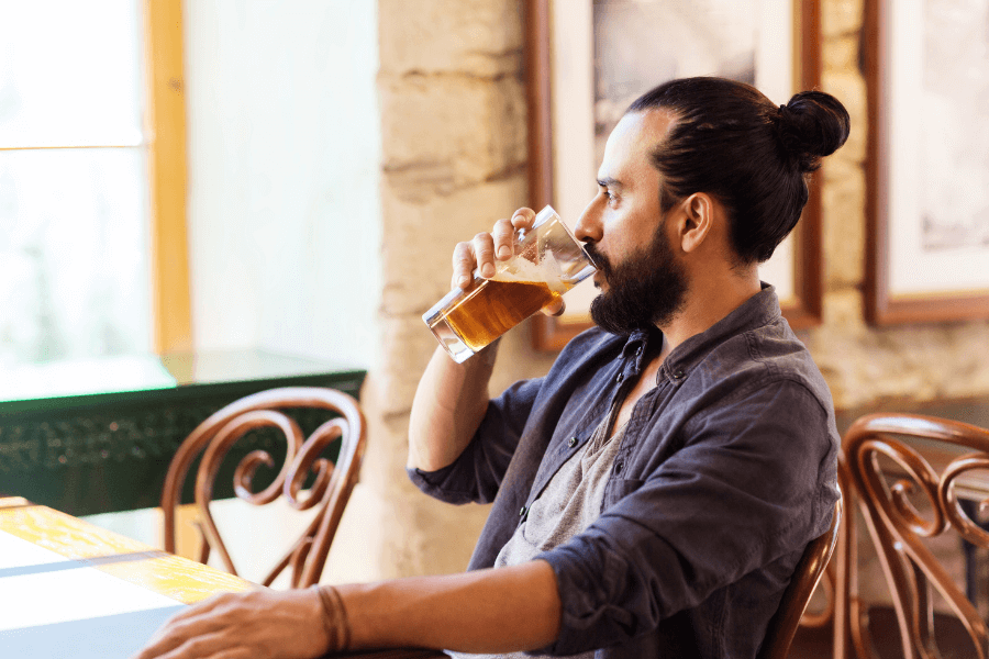 A man with a beard and hair tied in a bun sits in a rustic café or bar, drinking beer from a pint glass. The setting includes wooden furniture, stone walls, and framed artwork in the background.