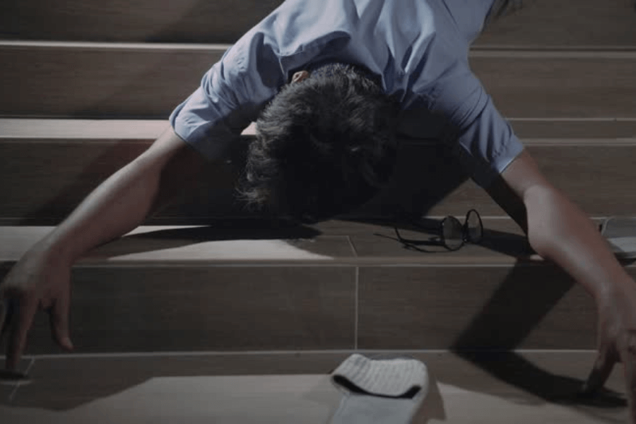 A man lies face down on a staircase, arms outstretched, with his glasses and a white object, possibly a piece of paper or cloth, scattered nearby. The dim lighting emphasizes a dramatic or somber mood.