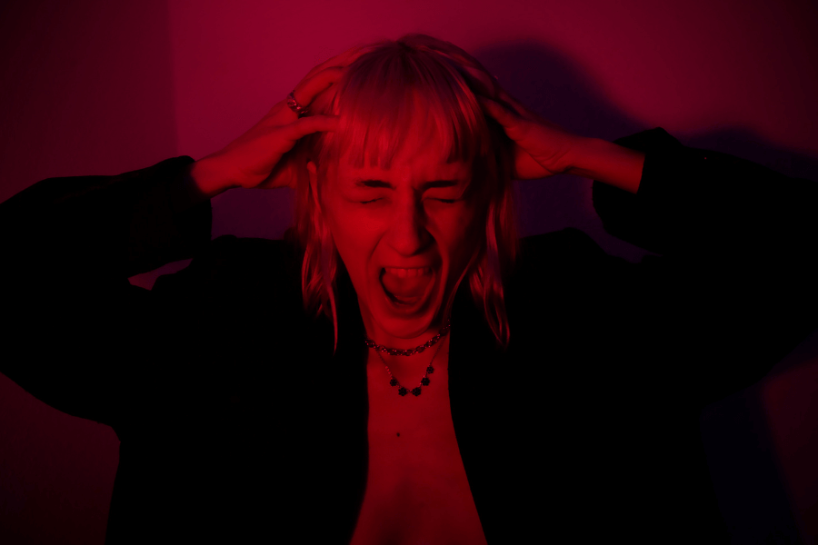 A woman with blonde hair and a black jacket holds her head with both hands and screams, her face expressing intense emotion. The dramatic red and purple lighting creates a moody, edgy atmosphere.