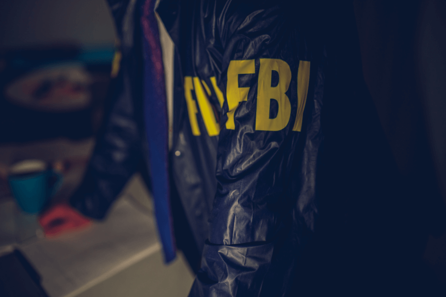 A close-up of a person wearing a dark jacket with "FBI" in large yellow letters on the sleeve, leaning over a table. The background is dimly lit, with a coffee cup and documents partially visible.