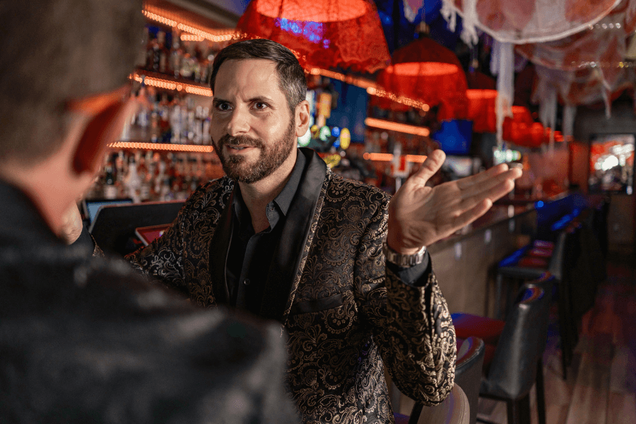 A man in a patterned blazer and black shirt gestures animatedly while speaking to someone at a bar. The background features red hanging lights, festive decorations, and a row of bar stools.