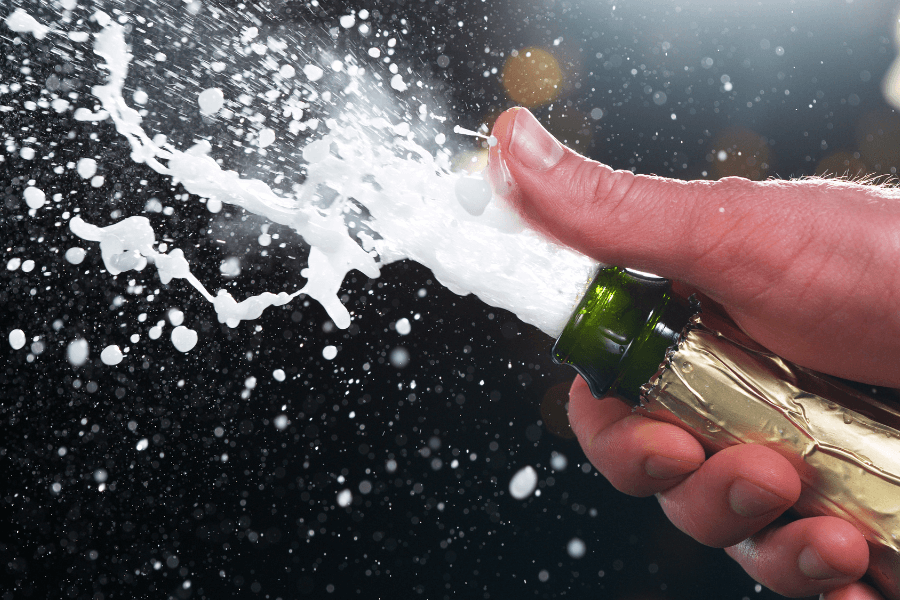 A close-up of a hand holding a champagne bottle as it is opened, with foam and liquid spraying out dramatically. The background is dark, highlighting the celebratory burst of bubbles.