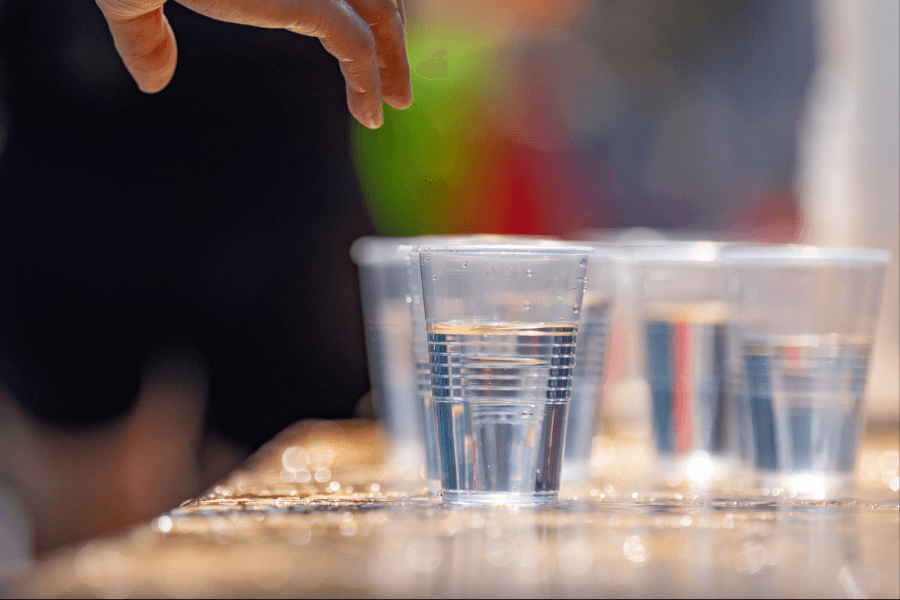 A hand reaches toward a row of clear plastic cups filled with water on a sunlit table. The background is blurred with bright, colorful shapes, suggesting an outdoor setting.