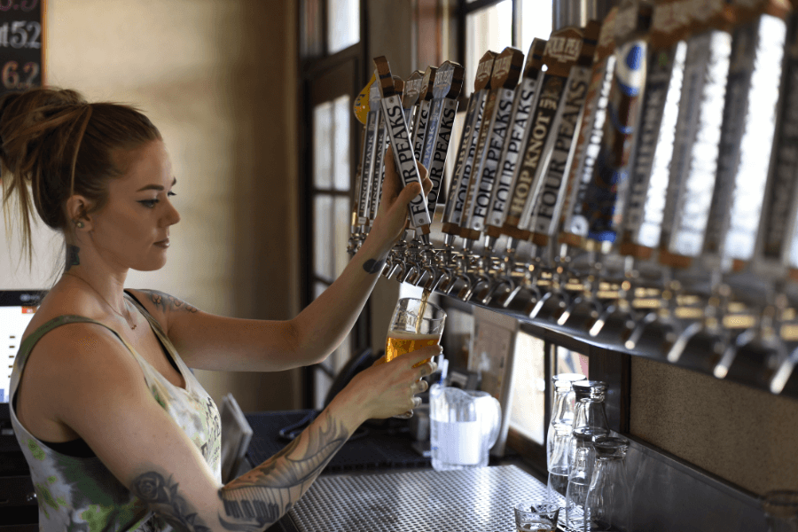 A bartender with tattoos and a tied-back hairstyle pours a draft beer from a row of labeled taps into a glass. The setting features natural light streaming through nearby windows and clean glassware on the counter.