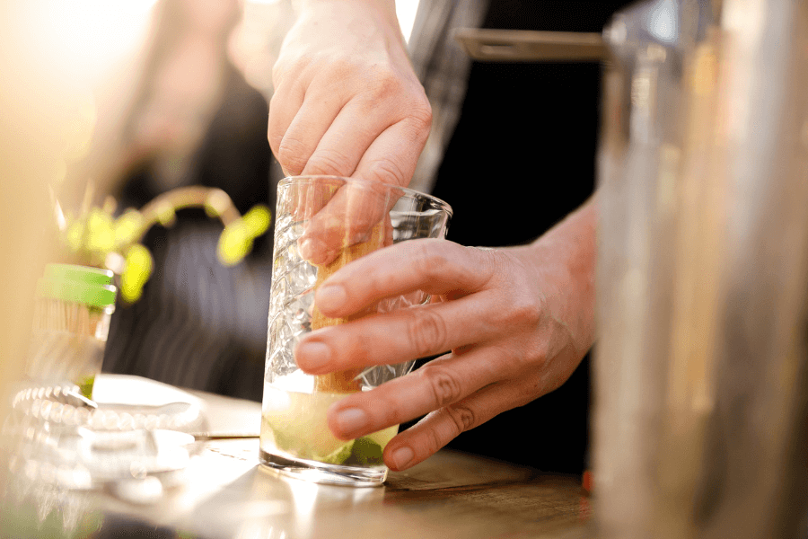 A close-up of hands using a wooden muddler to press lime wedges in a textured glass, preparing a cocktail. The background is blurred, suggesting a lively setting with plants and bartending tools.