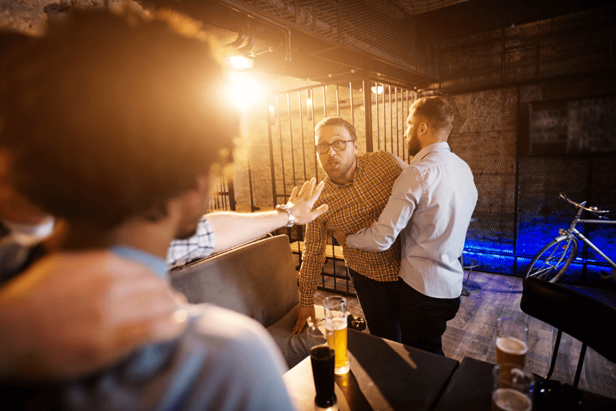  A tense moment in a bar as one man in a yellow plaid shirt is held back by another man, while gesturing and leaning toward someone off-screen. Beer glasses sit on the table in the foreground, and the background features a dimly lit industrial-style bar with a bicycle in view.