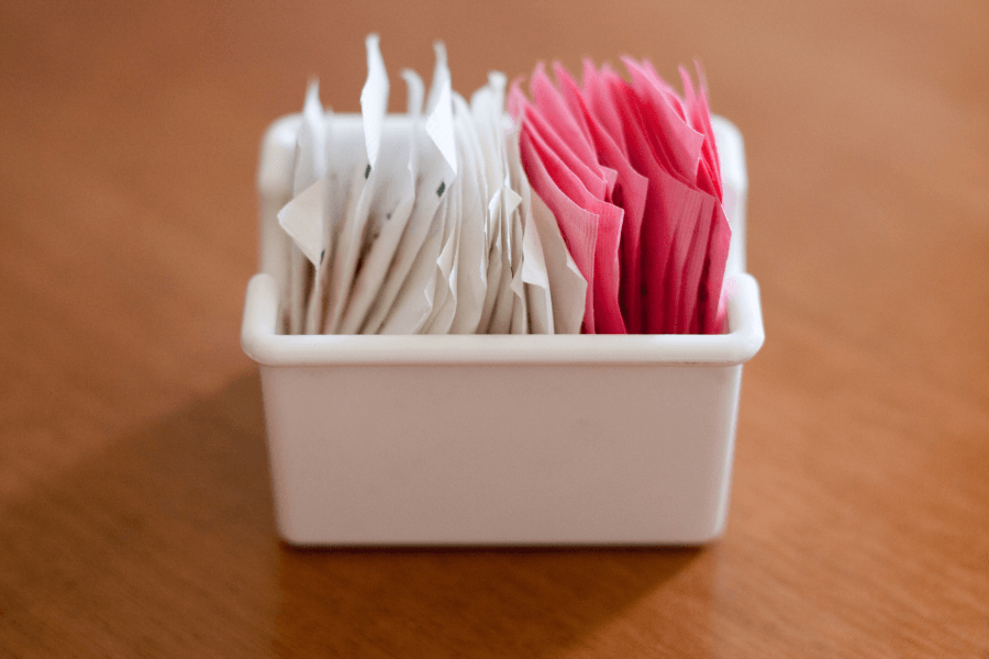 A small white container holding neatly arranged packets of sweeteners in white and pink wrappers sits on a wooden table. The scene is simple and clean, with a soft focus.