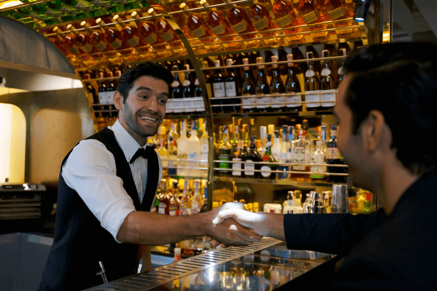 A bartender wearing a black vest and bow tie greets a customer with a handshake across a polished bar counter. The background showcases an ornate display of liquor bottles illuminated under warm lighting.