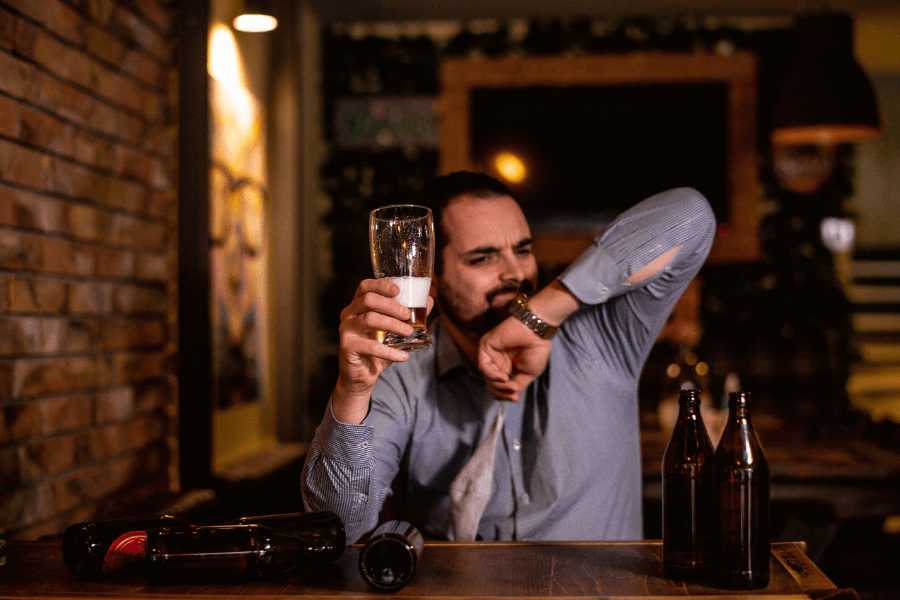 A man in a dress shirt sits at a bar table holding an almost-empty beer glass, wiping his face with his arm. Empty beer bottles are scattered on the wooden table, with a dimly lit, rustic bar in the background.