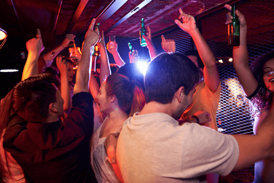 A lively crowd of people dancing and cheering in a nightclub, with hands raised holding drinks, including bottles and glasses. The dim red lighting and energetic atmosphere highlight the excitement of the celebration.