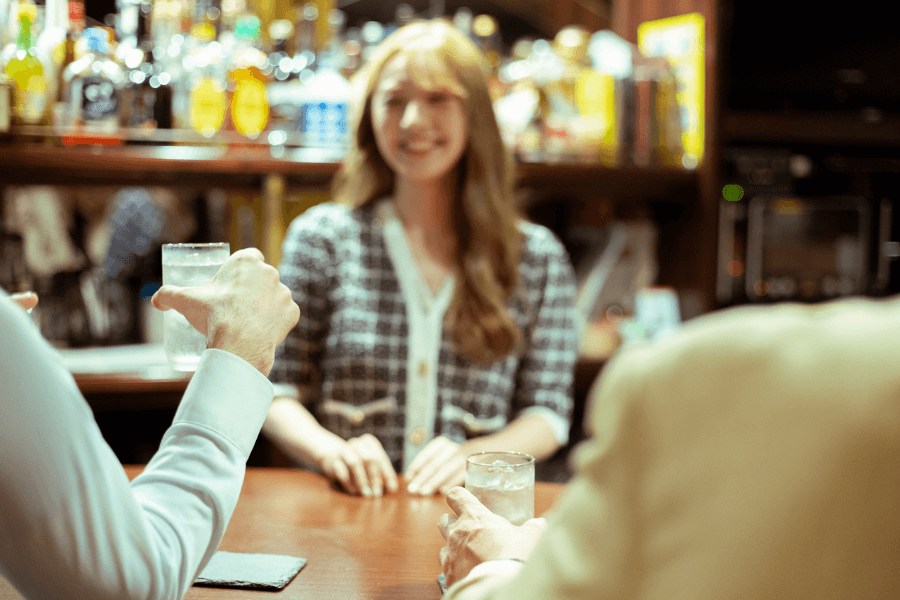 A smiling bartender with long, wavy hair stands behind the bar, engaging with two patrons holding glasses of clear drinks. The background is filled with shelves of bottles and a cozy, warmly lit atmosphere.