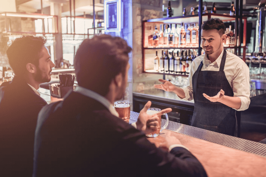 A bartender wearing an apron and white shirt gestures while speaking to two patrons sitting at the bar, each with a glass of beer. The modern bar setting is warmly lit, featuring shelves stocked with bottles and an industrial-style design.