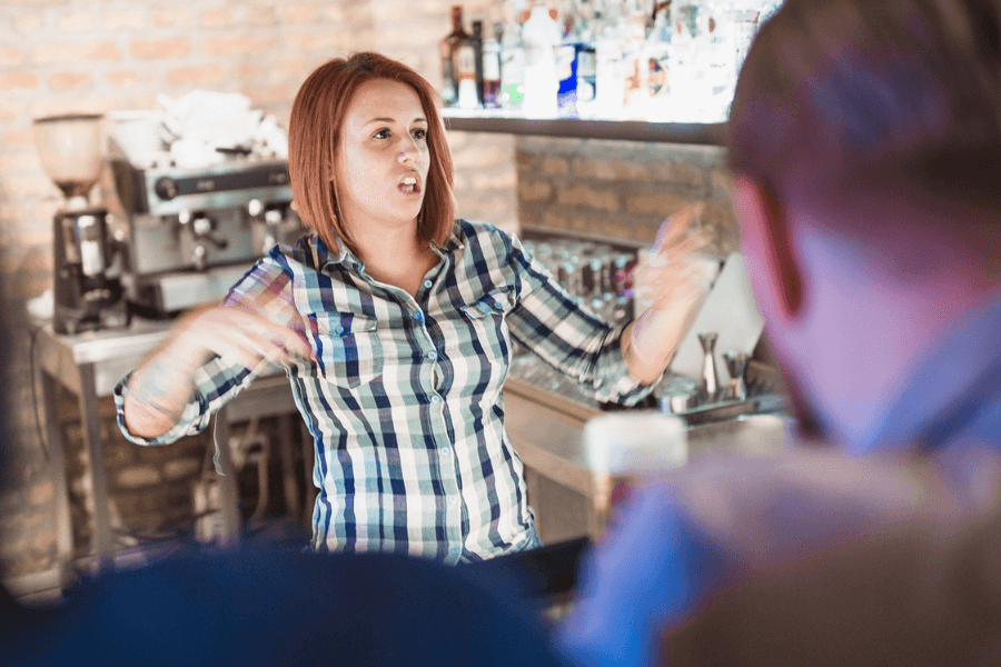 A bartender with red hair wearing a plaid shirt gestures animatedly while speaking to customers at the bar. The background shows a brick wall, shelves of liquor bottles, and an espresso machine in a cozy bar setting.