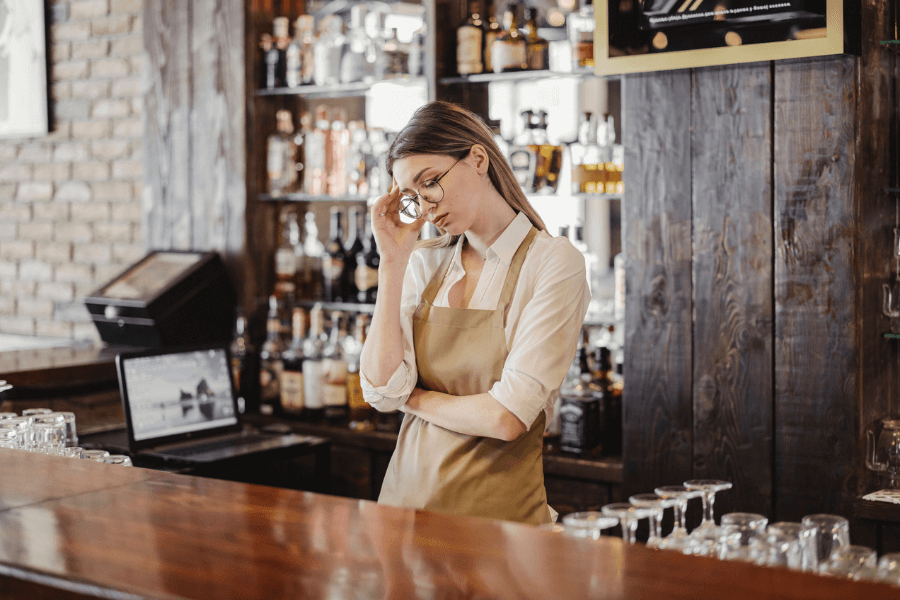 A bartender wearing an apron and glasses stands behind a wooden bar, holding her temple with a thoughtful or stressed expression. The background features shelves filled with bottles of alcohol and a laptop on the counter.