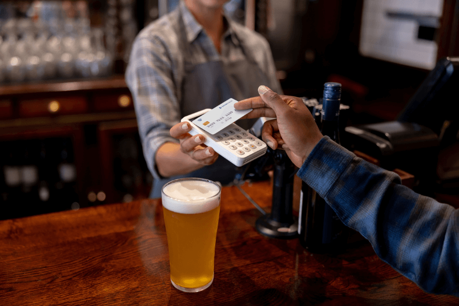 A bartender wearing an apron holds out a payment terminal while a customer taps a credit card to pay. A frothy glass of beer sits on the polished wooden bar counter in a warmly lit setting.