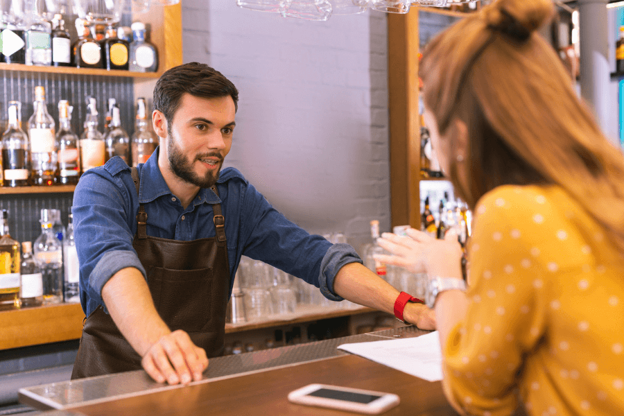 A bartender wearing a leather apron and denim shirt leans on the bar counter while attentively speaking to a customer. The customer gestures animatedly, and a smartphone lies on the counter in a modern bar with a well-stocked shelf of bottles in the background.