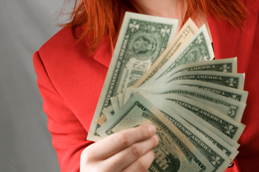 A person with vibrant red hair wearing a red jacket holds a fan of US dollar bills in their hand, prominently displaying the currency. The focus is on the money, with the person's face out of frame.