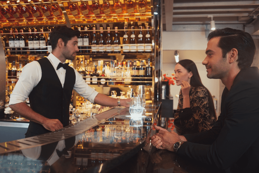 A bartender in a black vest and bow tie serves drinks to a man and a woman seated at a sleek bar. The bar is lined with bottles of alcohol displayed on shelves behind the bartender, creating a luxurious and upscale ambiance.