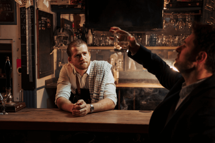 A bartender with a towel over his shoulder leans on a wooden bar counter, listening attentively to a male customer holding up a glass of whiskey. The setting is dimly lit with shelves of glassware and bottles in the background, giving the scene a cozy pub vibe.