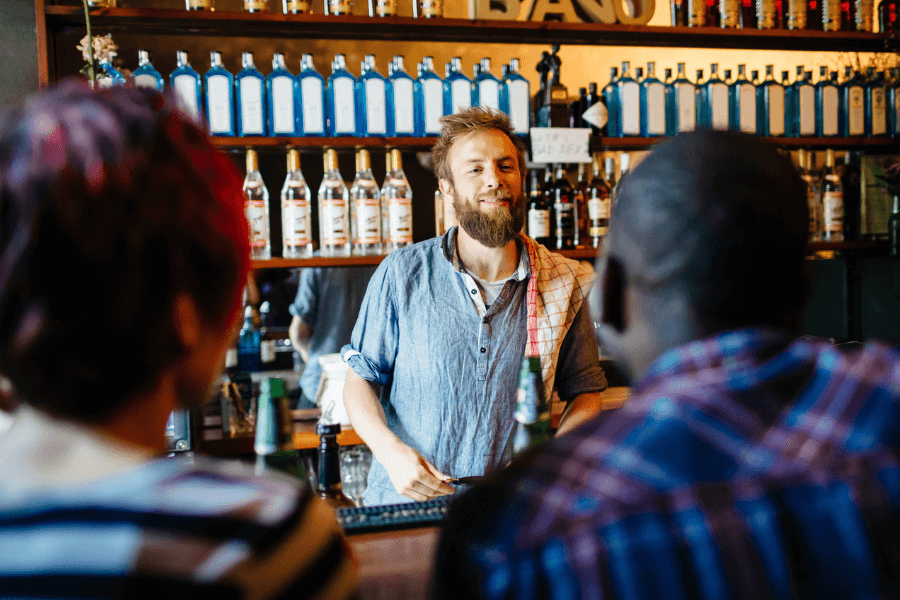 A bearded bartender in a casual button-up shirt smiles while interacting with two patrons sitting at the bar. A towel is draped over his shoulder, and shelves stocked with an array of liquor bottles are neatly organized in the background, creating a relaxed and inviting atmosphere.