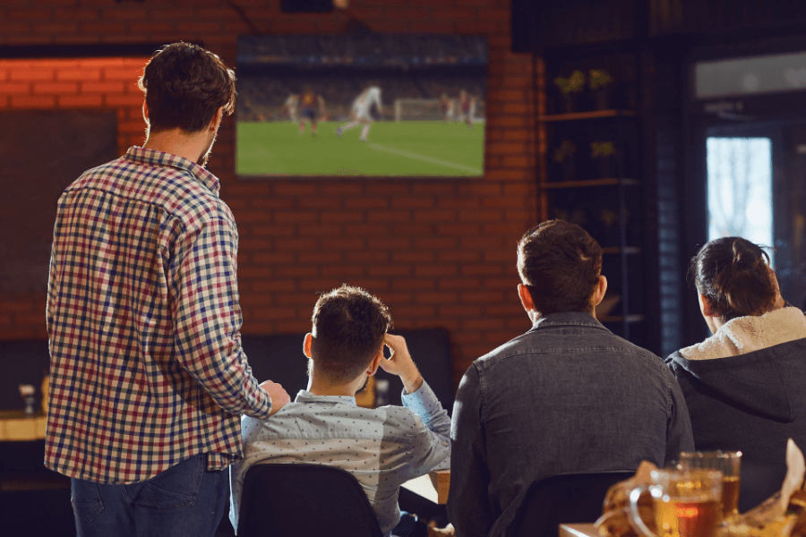 A group of four people sits and stands in a bar or lounge, watching a soccer game on a wall-mounted TV. The setting has brick walls, warm lighting, and beer glasses visible on the table in the foreground.