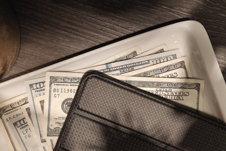 A stack of hundred-dollar bills is placed in a white dish, partially covered by a textured gray wallet. The surface underneath is a wooden table with soft lighting.