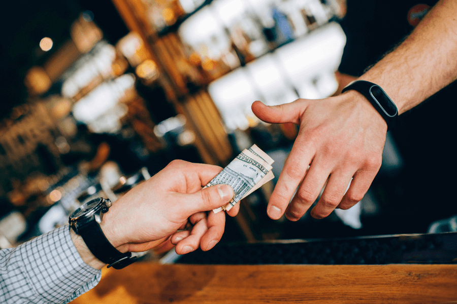 A close-up of two hands exchanging money over a bar counter; one hand holds a folded bill, passing it to the other. The background is blurred, showing shelves of bottles and a bar setting.