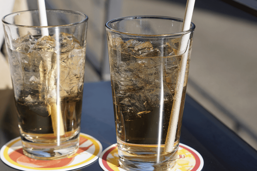 Two tall glasses filled with ice and a light golden drink, each with a white straw, placed on colorful coasters on a dark table. The setting is outdoors with sunlight reflecting off the glasses.