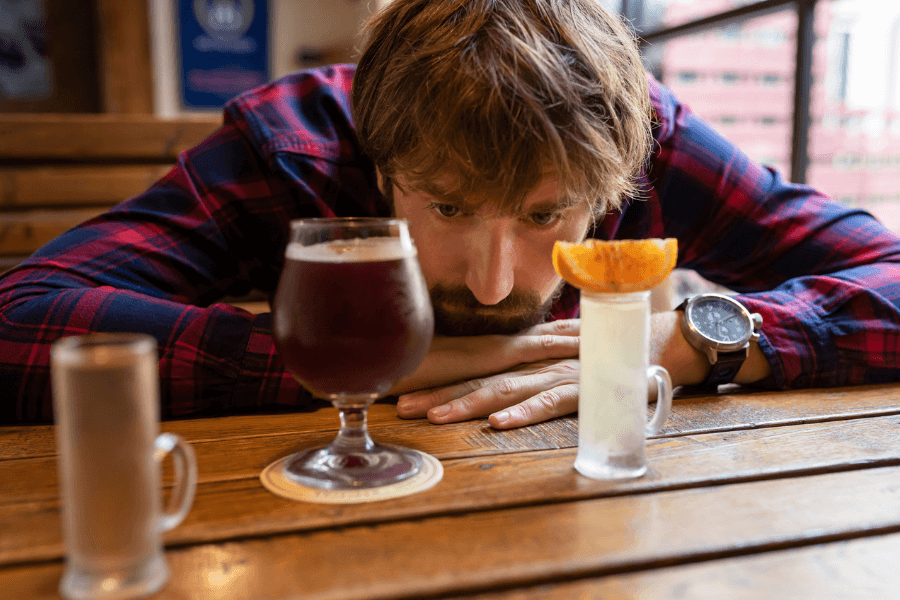 A man in a red plaid shirt rests his head on a wooden table, staring intently at three drinks in front of him: a dark beer in a goblet, a clear cocktail garnished with an orange slice, and a shot glass. The setting appears to be a casual bar with wooden furniture and urban views in the background.