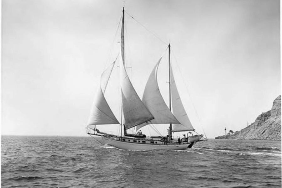 Black-and-white photo of a large sailboat with full sails cutting through open water, moving past a rocky shoreline with a lighthouse in the distance. The vessel has two masts and several people aboard, visible near the helm and along the deck.