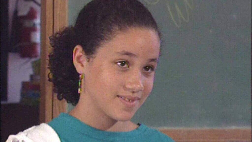 A young girl with curly hair pulled back smiles softly while standing in front of a chalkboard. She wears colorful dangling earrings and a teal shirt, exuding a calm and thoughtful expression.