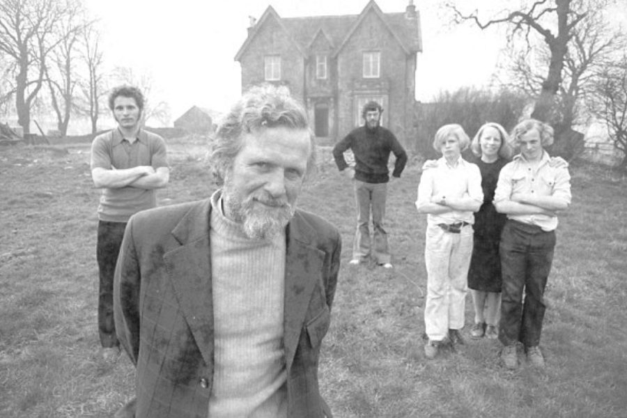 Black-and-white photo of a group of seven people standing on a grassy field in front of a large, old stone house with steep gables. The bearded man in the foreground wears a turtleneck and blazer, while the others stand further back with arms crossed, some smiling, surrounded by bare trees and a misty backdrop.
