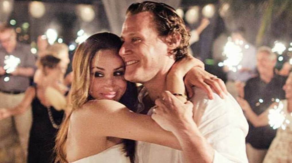 A woman in a white dress embraces a smiling man at a party or celebration. They are surrounded by people holding sparklers, creating a festive and romantic atmosphere.