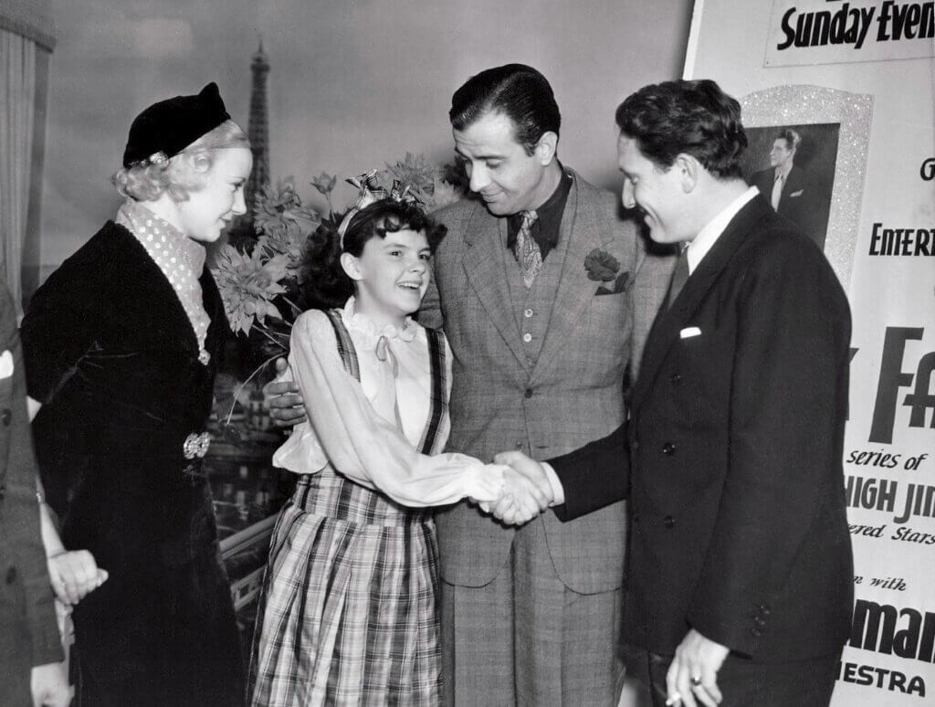 Black-and-white photo of a young girl in a plaid dress shaking hands with a smiling man in a suit. Two other adults, a man in a checkered three-piece suit and a woman in a dark dress with a hat, look on with cheerful expressions; a large poster and the Eiffel Tower are visible in the background.