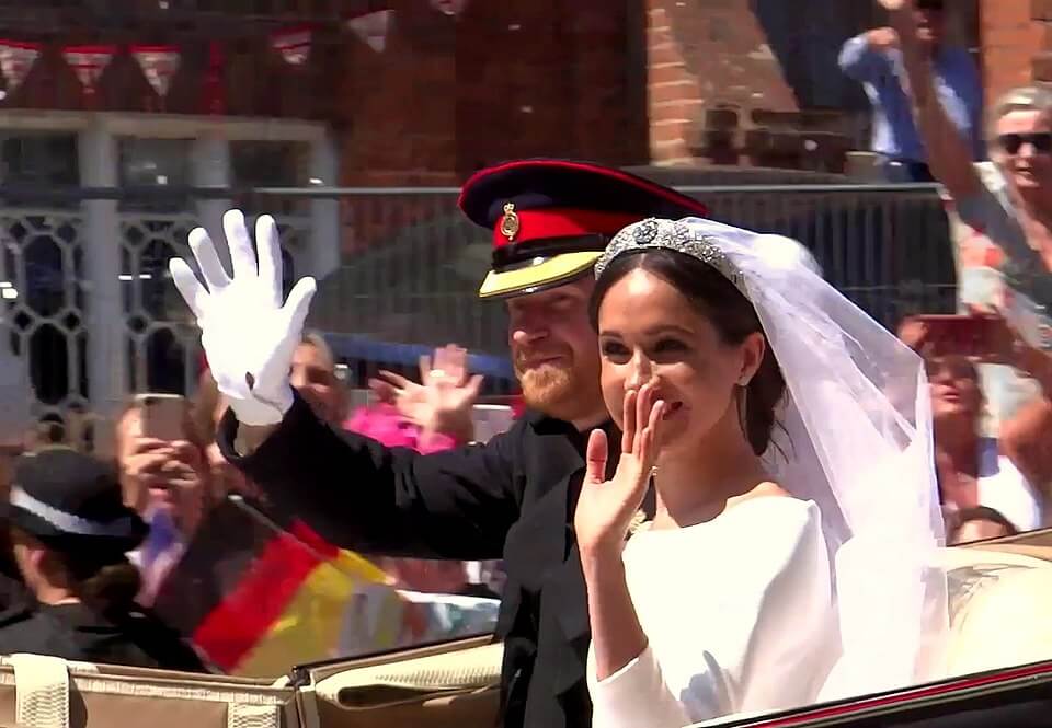 A bride in a white gown and veil waves alongside a man in a military uniform during a public procession. They are riding in an open carriage, surrounded by cheering crowds and waving flags.