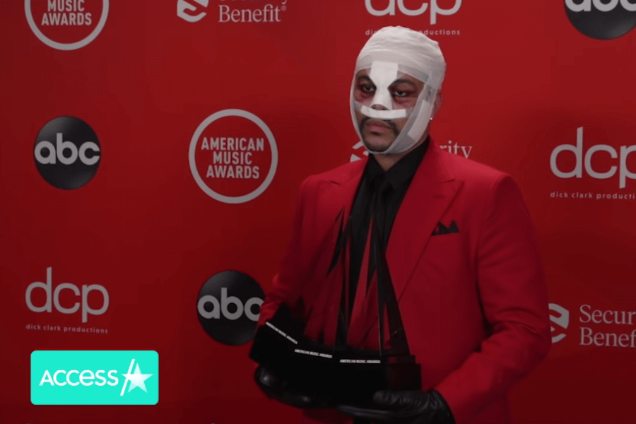 The Weeknd stands in front of an American Music Awards backdrop wearing a red suit, black shirt and gloves, and full face bandages with bruised makeup underneath, holding multiple AMA trophies. His appearance is part of a visual statement tied to his “After Hours” album era, reflecting themes of fame and physical transformation.