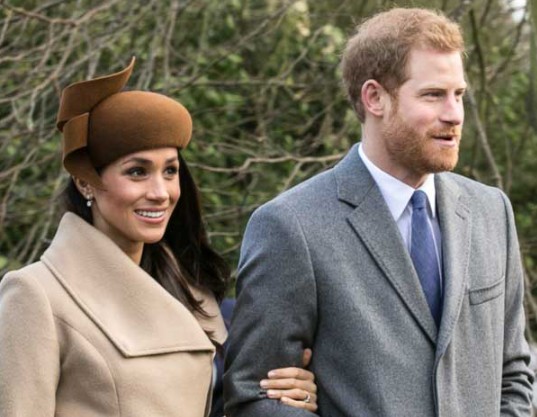 A smiling woman in a tan coat and brown hat walks arm-in-arm with a man in a gray suit and blue tie. They are outdoors, surrounded by trees, and appear to be at a formal event.