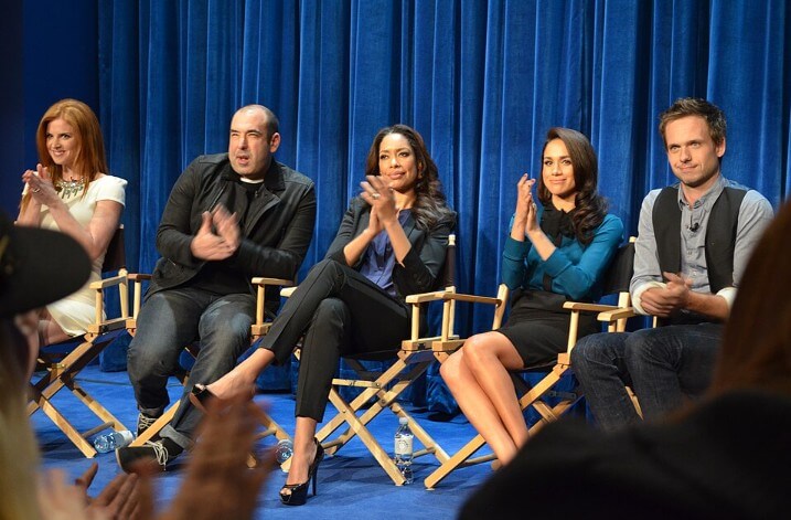 Five actors sit on director's chairs in front of a royal blue curtain, applauding toward an unseen audience. They appear to be part of a panel or cast event, dressed in smart casual and formal attire, with water bottles at their feet.