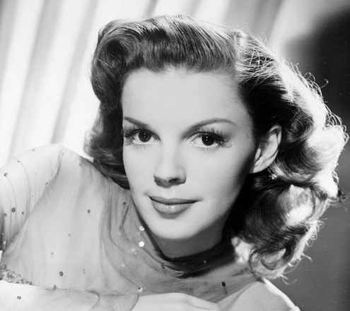 Black and white portrait of a woman with wavy shoulder-length hair, styled in 1940s fashion. She has a soft smile and is wearing a sheer, sparkly high-neck top, gazing directly at the camera with a composed expression.