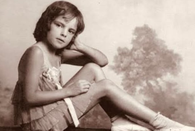 Sepia-toned photograph of a young girl in a ballet outfit sitting sideways on a ledge. She wears ballet slippers and a ruffled dress, resting her head on one hand with a calm, poised expression.